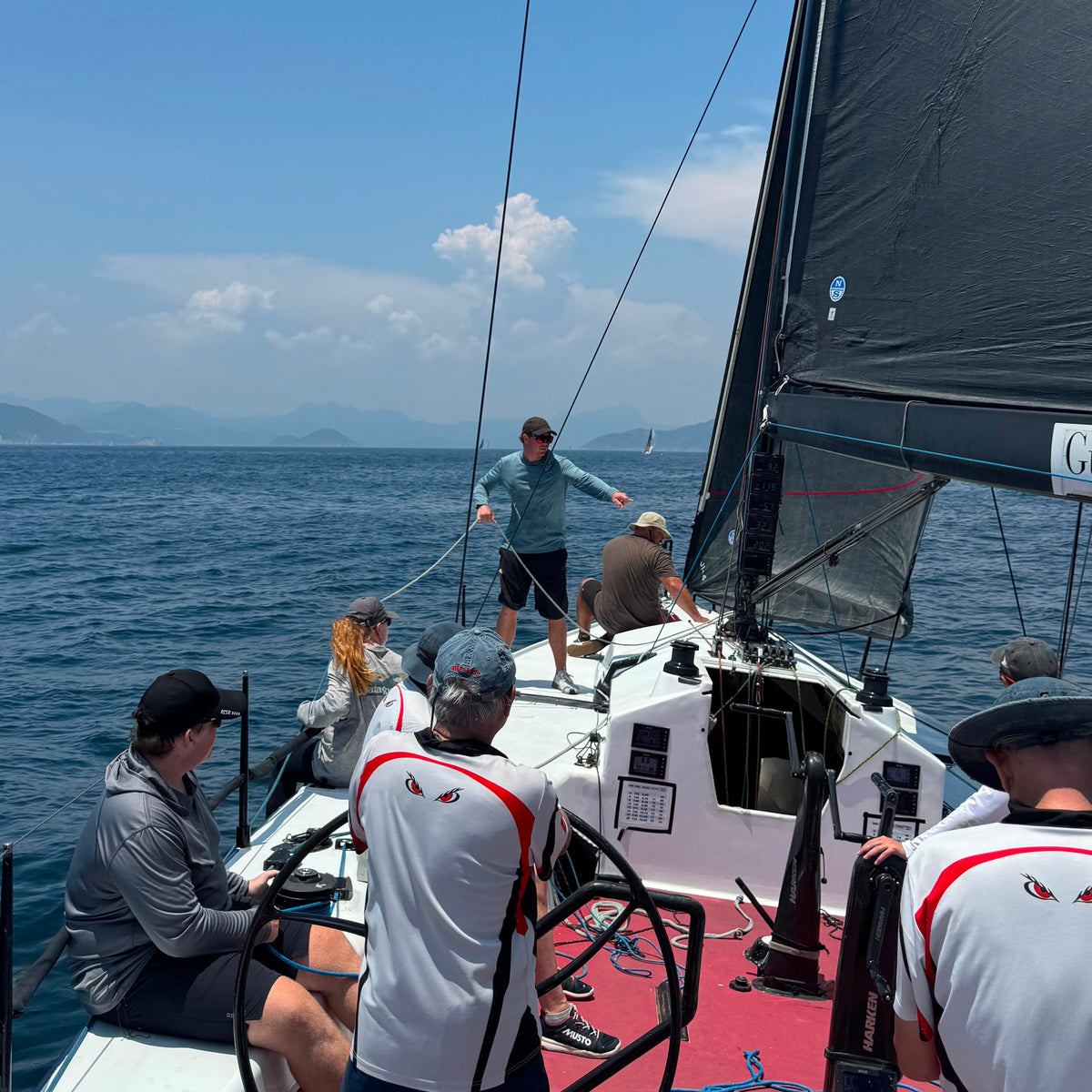 A group of sailors on a white sailboat with a black mainsail, sailing on open blue water under a clear blue sky.