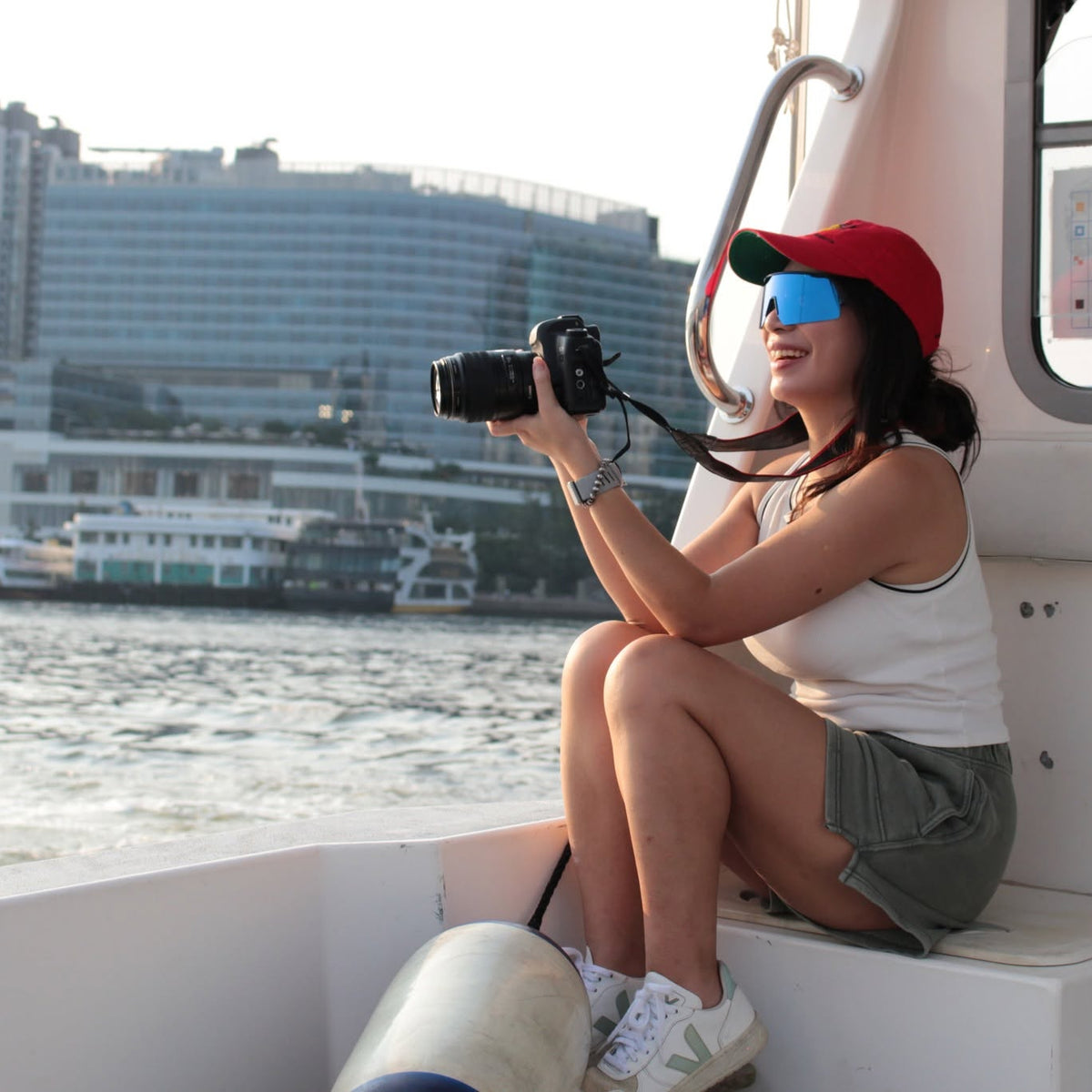 A woman sitting on the white deck of a boat, holding a DSLR camera. She is wearing a red baseball cap, blue-tinted sunglasses, a white tank top, and olive green shorts, with a waterfront city skyline in the background.