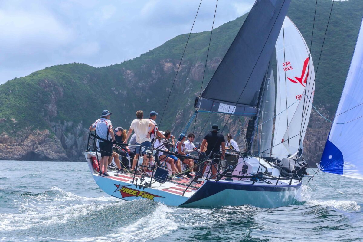 A racing sailboat with multiple crew members on board racing on the open sea, with a green mountainous coastline in the background under a cloudy sky.