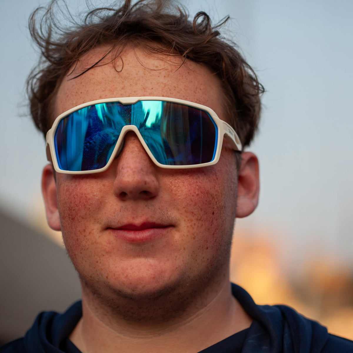 A close-up portrait of a man wearing oversized shield-style sunglasses with a white frame and reflective blue mirrored lenses, set against a softly blurred outdoor background.